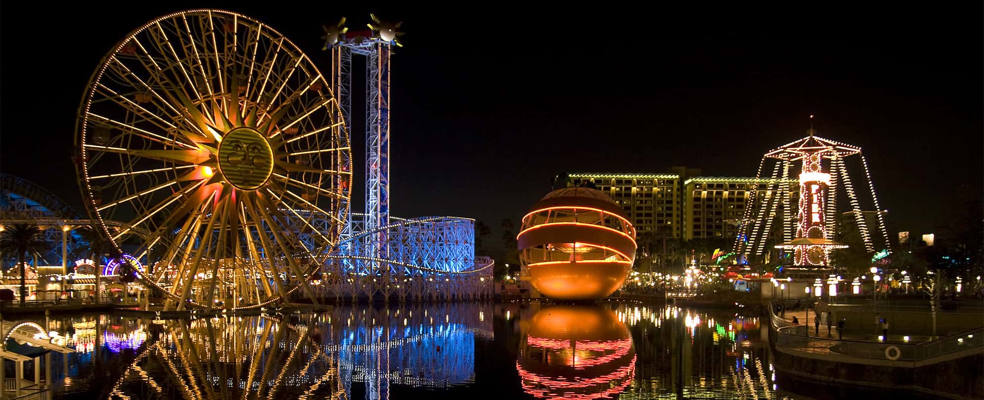 Disney's California Adventure theme park with a Ferris wheel and rides lit up at night and reflecting on the water.