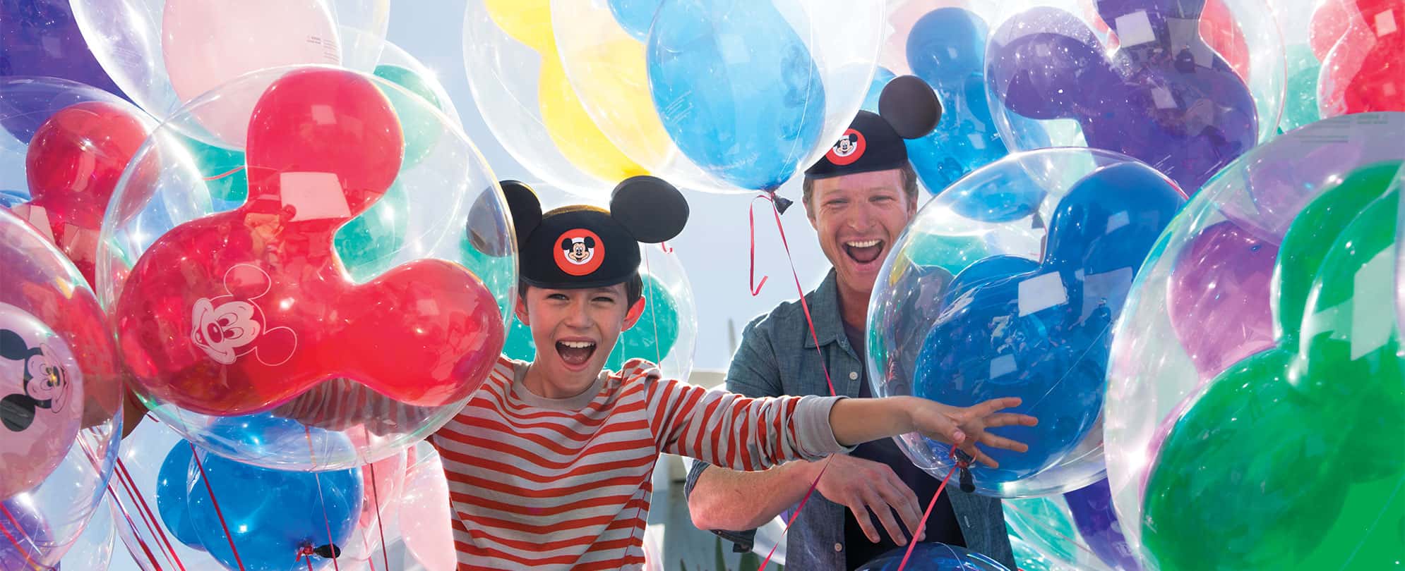 A kid and his dad wearing Mickey Mouse hats surrounded by colorful balloons at Disneyland.