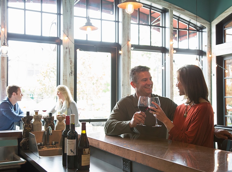 A couple sitting at a bar, both toasting to each other with a glass of red wine. 