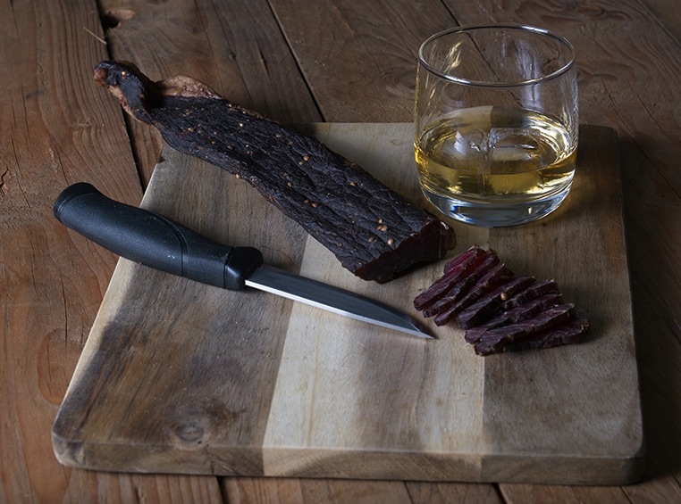 A wooden chopping block with a glass of whiskey, a knife, and freshly cut slices of salami on top of it. 