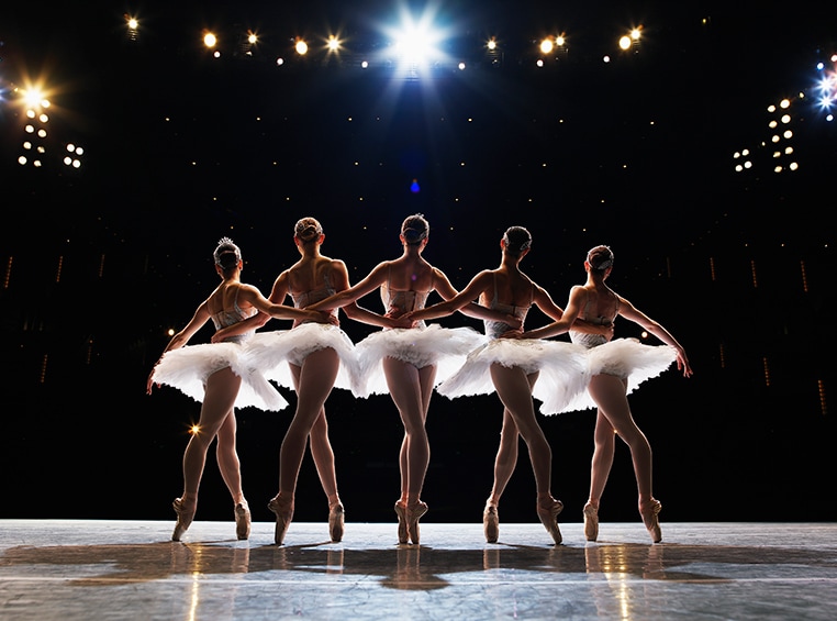 Five female ballerinas on stage holding each other waists in relever pose. 