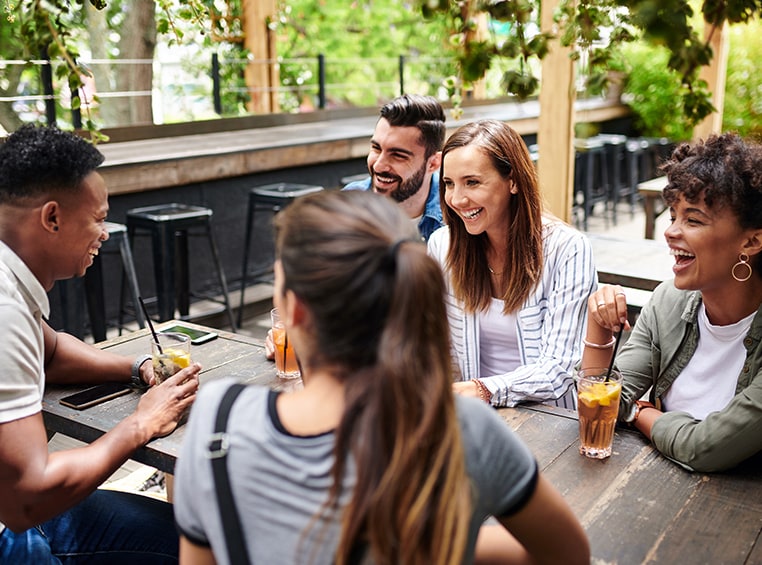 A group of five friends, sitting together at a table, socializing, enjoying a drink, and a good laugh. 