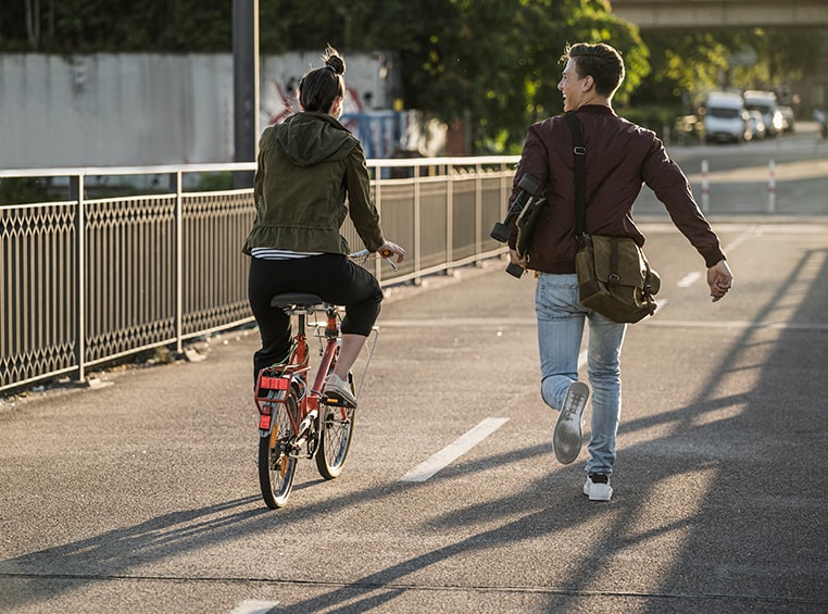 A caucasian male holding a skateboard and a caucasian female riding a bike, on an urban path.
