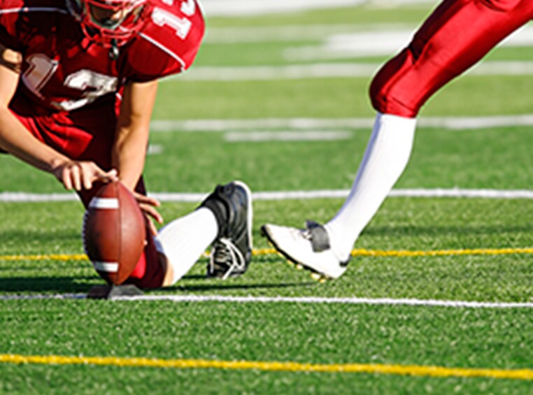A football player with his hand setting the ball as the other player prepares to kick a field goal. 