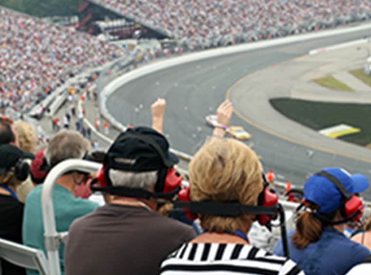 A caucasian couple are watching a car race at the race track with headphones on.