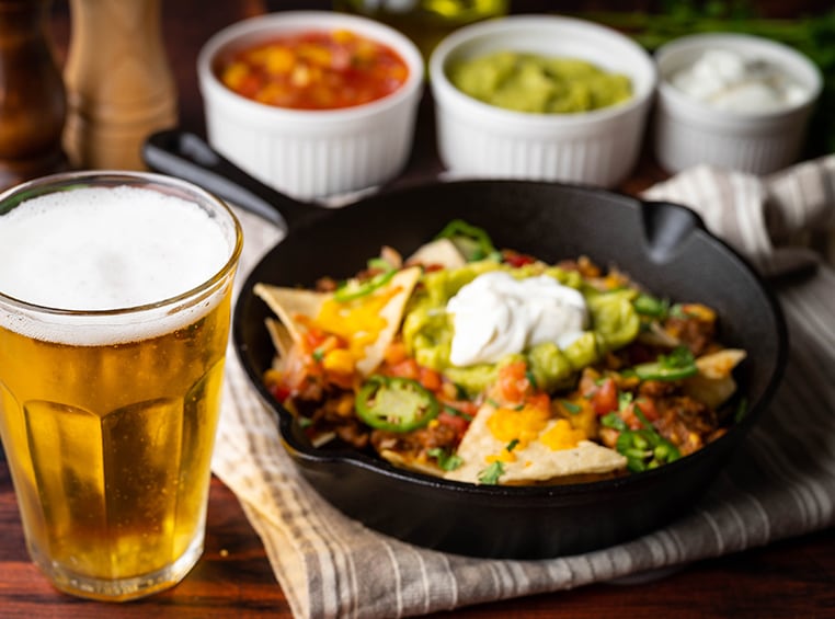 A tall glass of beer on a table, next to a skillet of nachos, salsa, guacamole, and sour cream. 