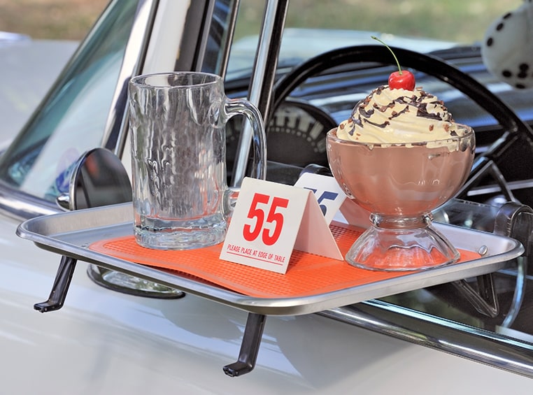 A drive-in food tray sits on a car window, with a milkshake on it.