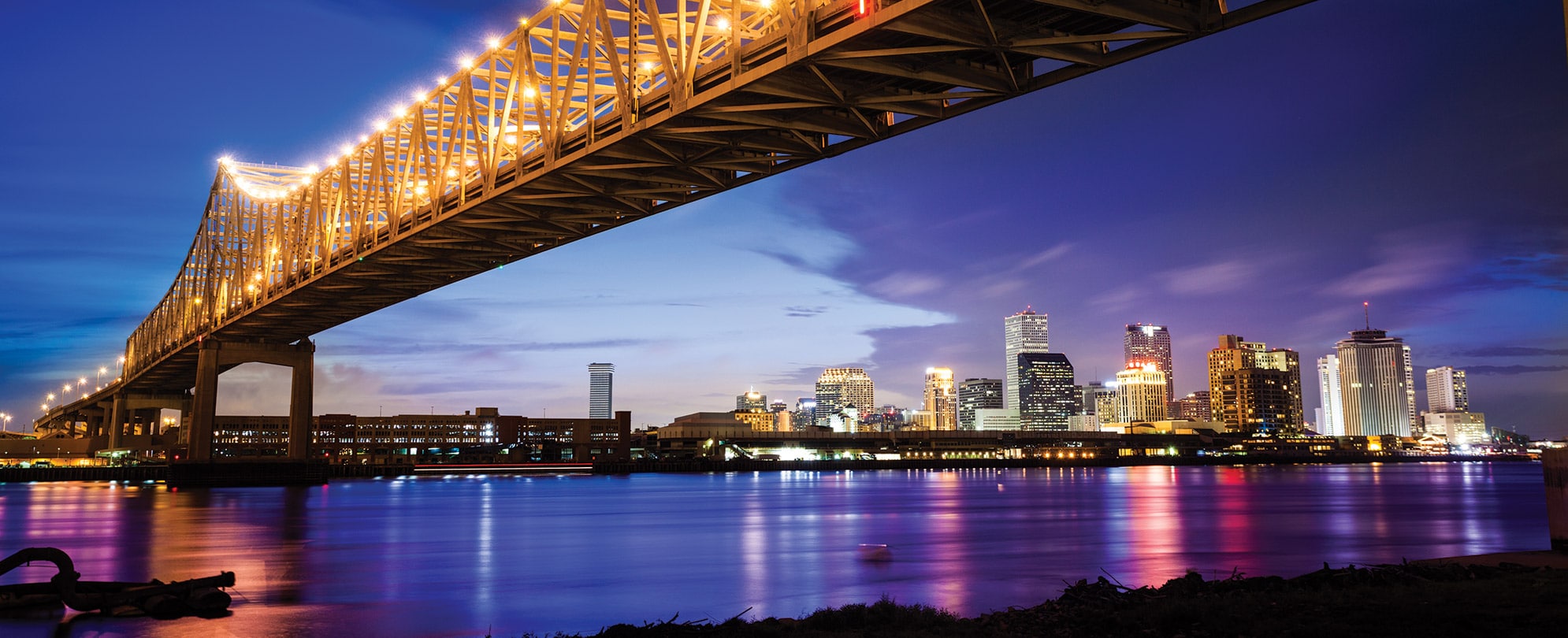 A river and bridge in New Orleans at night, the city skyline lit up in the distance.