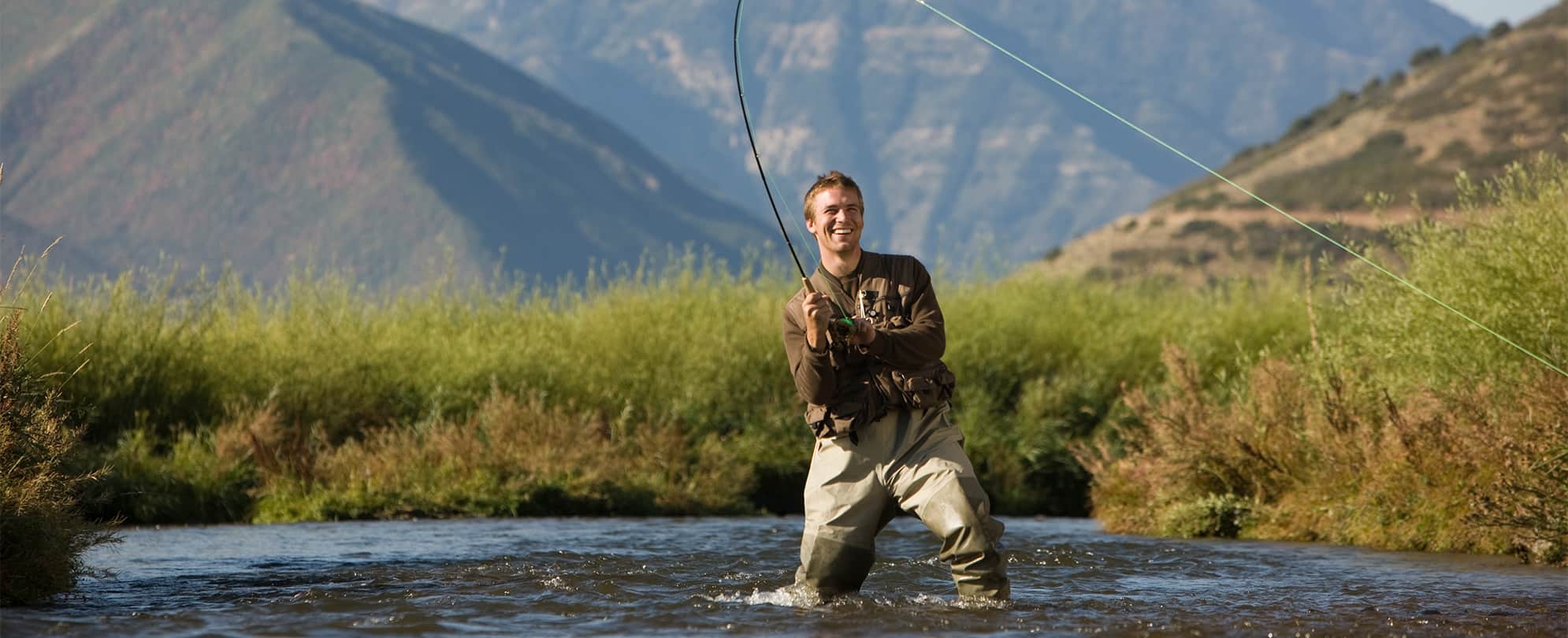 A smiling man fishing in a river with mountains in the background in Park City, Utah.