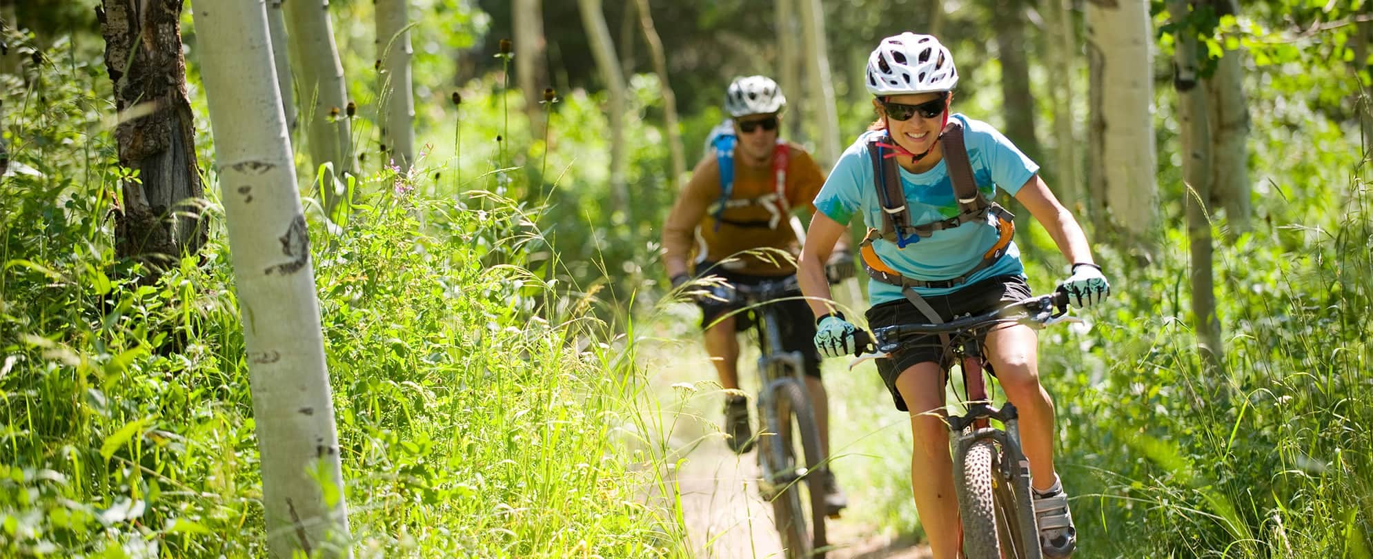 A man and woman wearing helmets and sunglasses ride bikes along a trail in Park City, Utah.