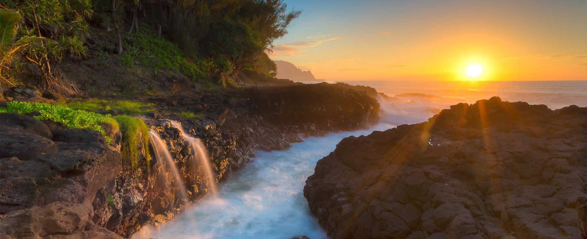 The sun sets over the ocean shining on the rocky coast of Princeville, Kauai in Hawaii