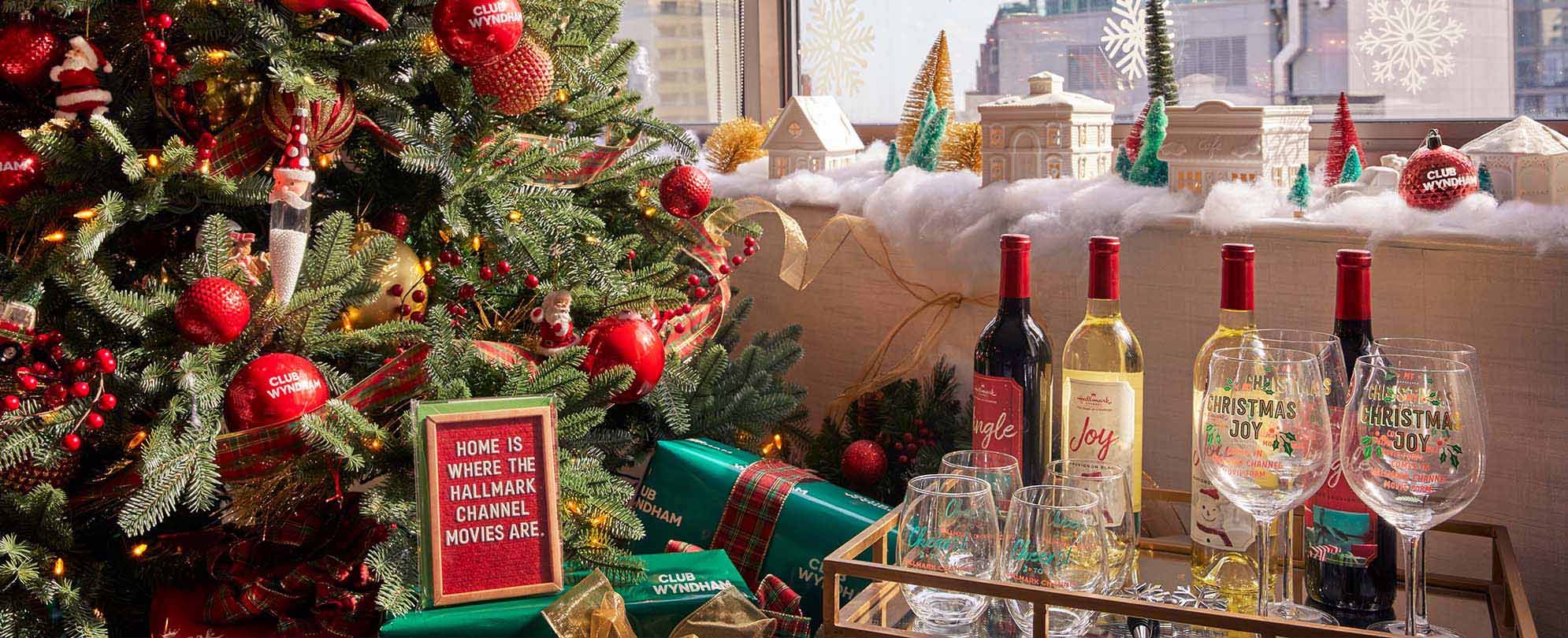 A bar cart with four holiday wines and Christmas decorated wine glasses next to the Christmas tree and a sign that says, "Home is where the Hallmark Channel movies are."