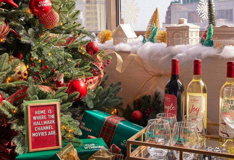 A bar cart with four holiday wines and Christmas decorated wine glasses next to the Christmas tree and a sign that says, "Home is where the Hallmark Channel movies are."
