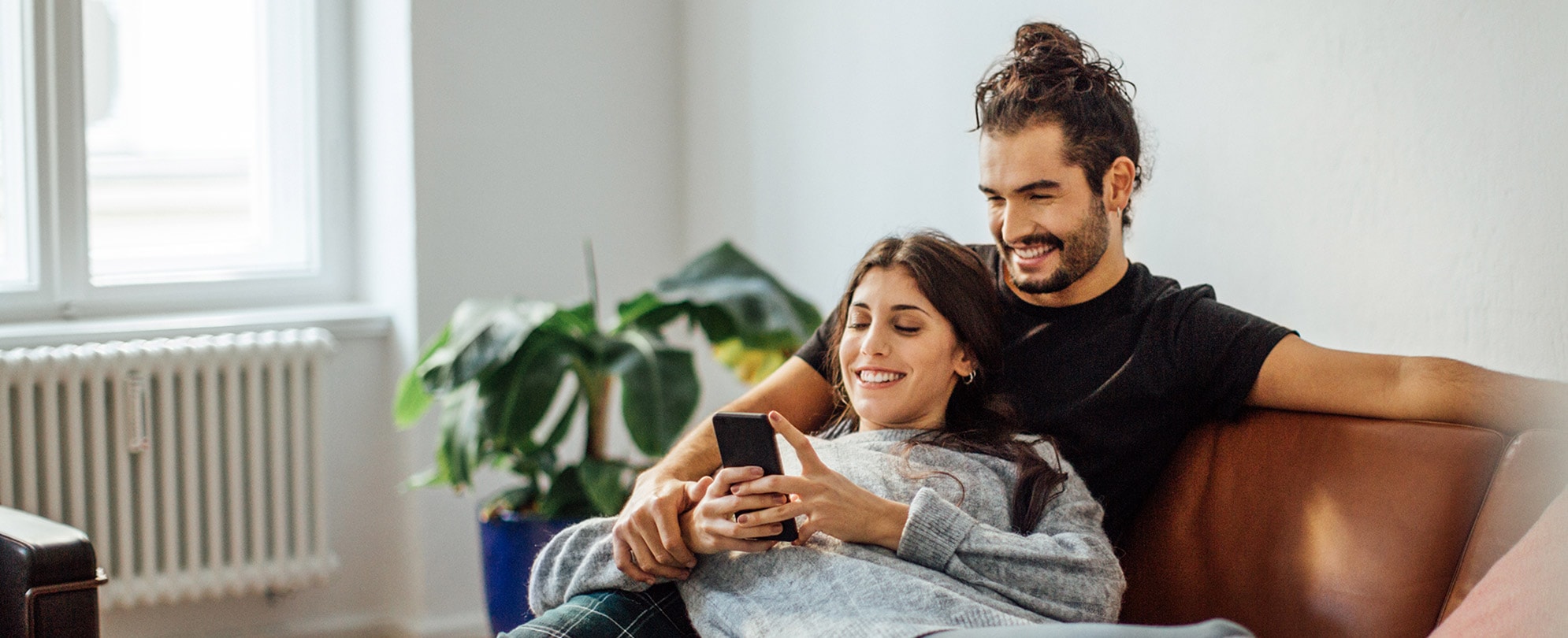 A man and woman sitting on a sofa together, smiling while looking at a cell phone.