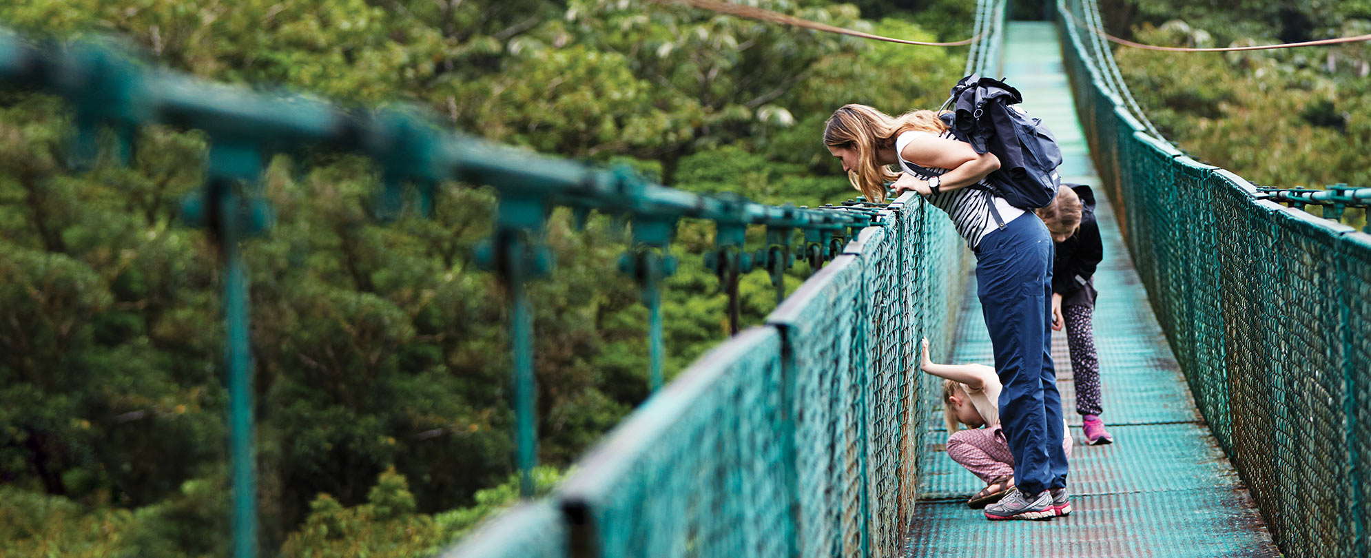 Two kids and a woman wearing a backpack look down over a suspended bridge.