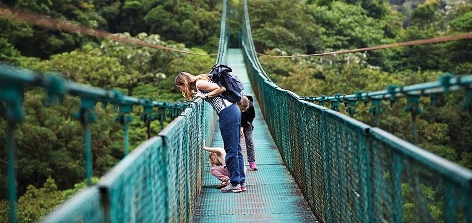 Two kids and a woman wearing a backpack look down over a suspended bridge.