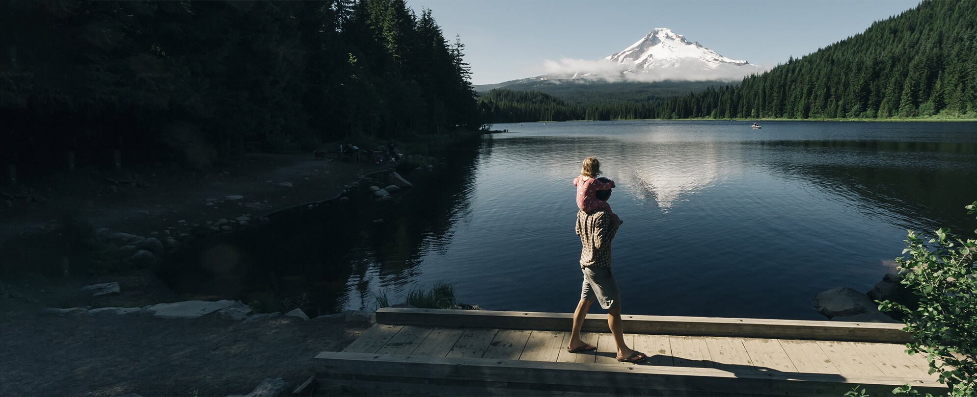 A dad holds a child on his shoulders in front of a Pacific Northwest lake with a mountain in the distance.