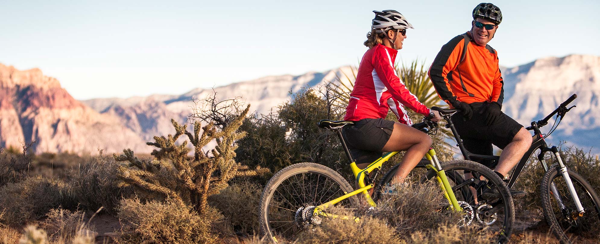 A man and woman smiling on bikes while surrounded by greenery and mountains. 