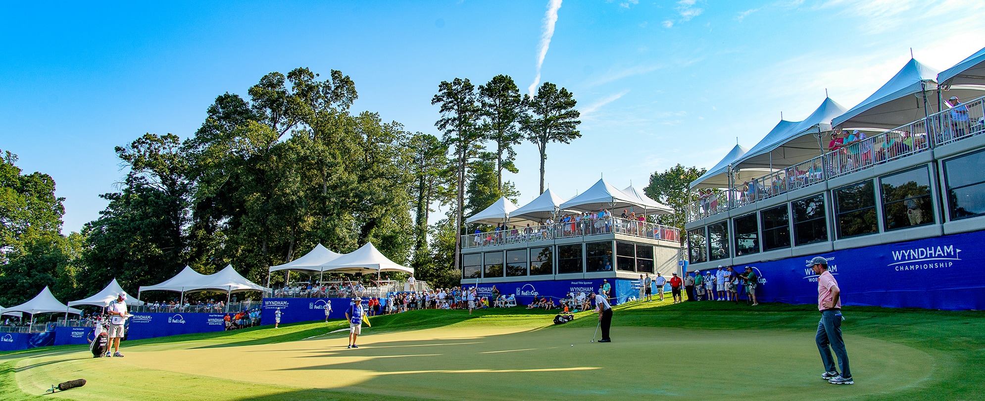Golfers on a green with spectators watching from under tents at the Wyndham Championship.
