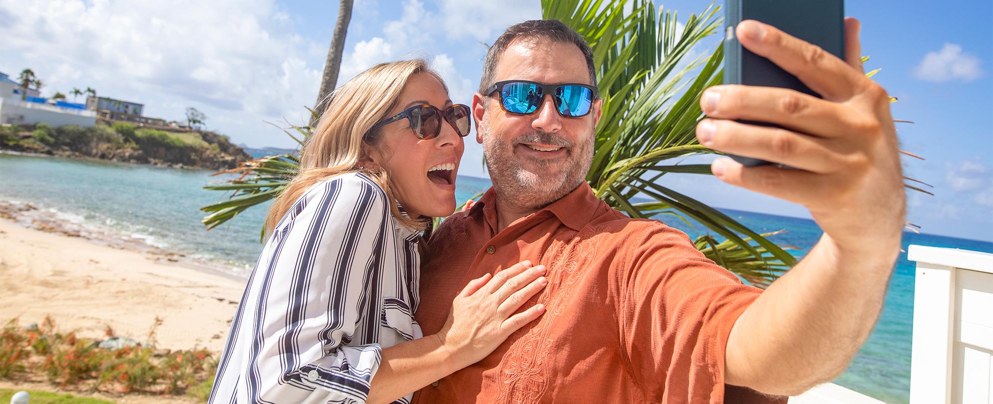 A man in sunglasses holding his phone out smiling, as a woman in sunglasses with her hand on his chest is smiling ready for him to take a photo on the beach with a palm tree and the ocean behind them. 