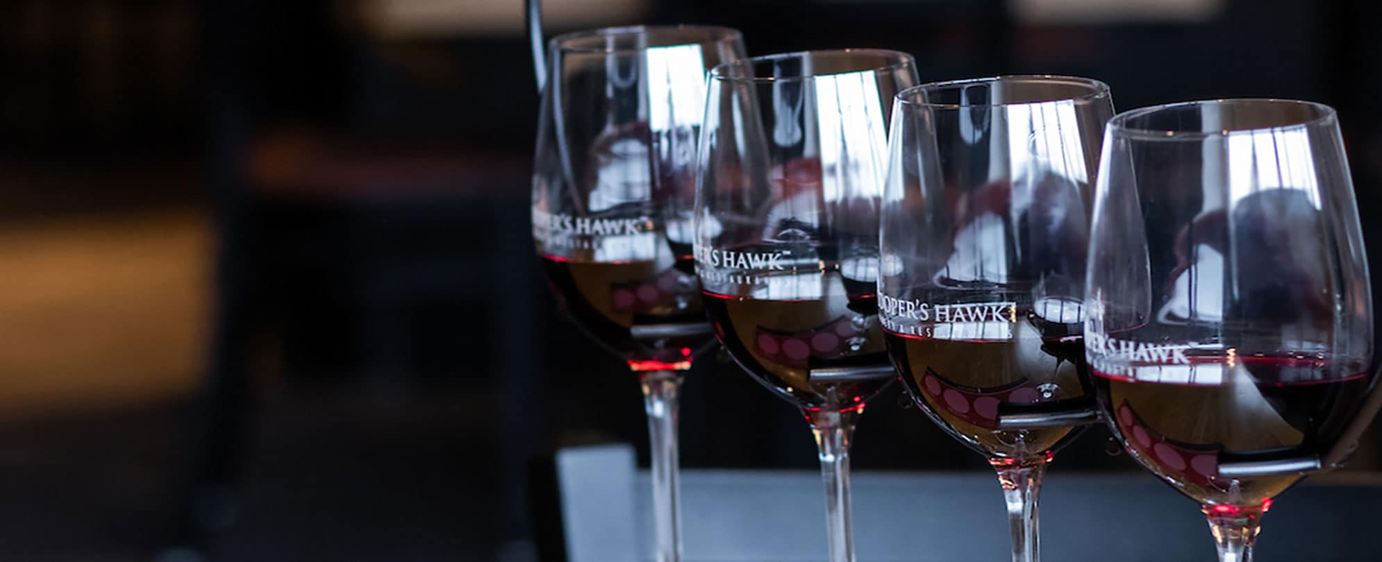 A row of four Cooper’s Hawk wine glasses filled with red wine.