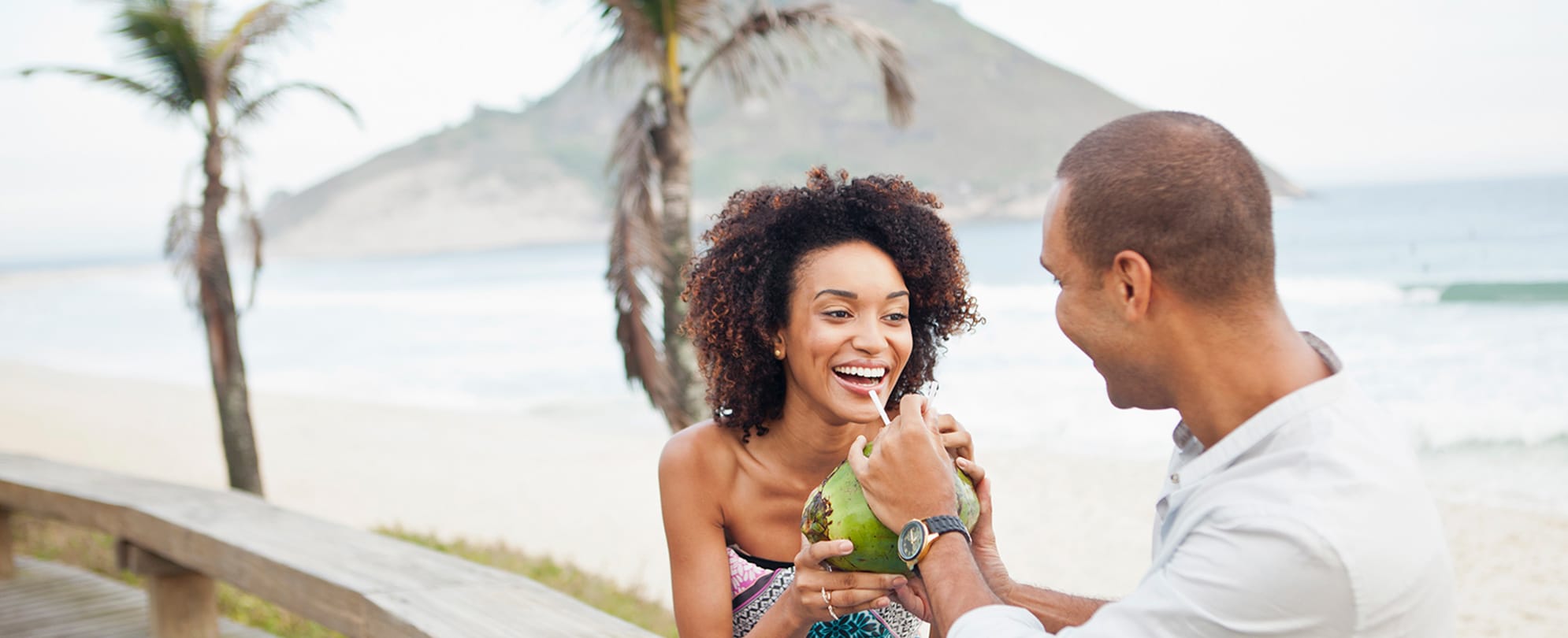 A man is holding a straw inside a coconut for the woman standing in front of him to take a sip of the coconut water. 