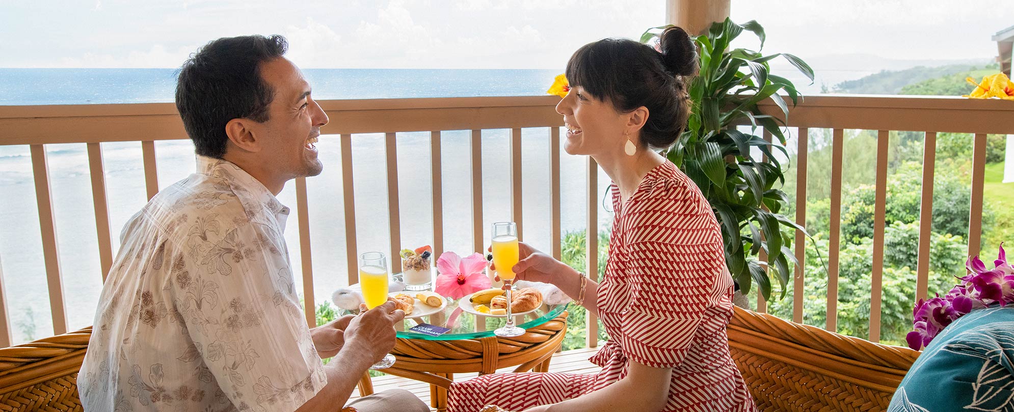A couple enjoys mimosas and pastries on their resort lanai, the ocean visible in the distance.