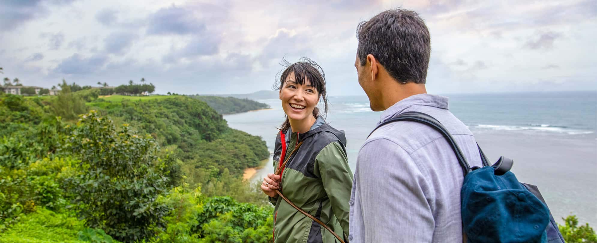 A woman turns around smiling at a man as they hike along a lush ridge with the ocean in the distance.