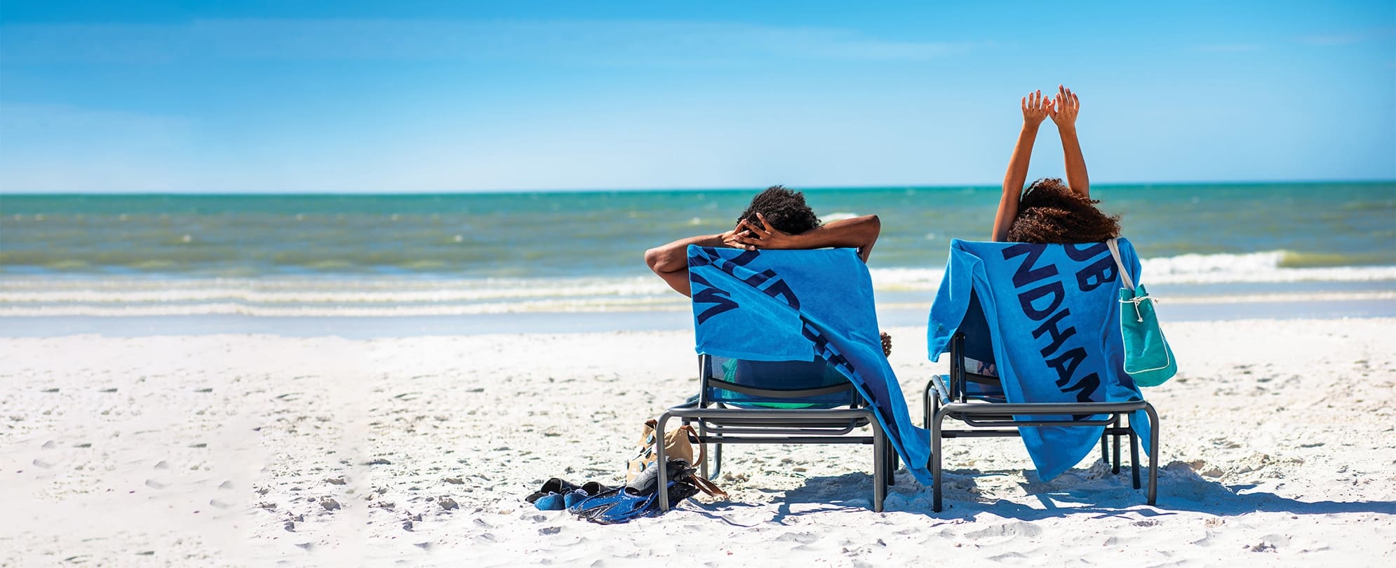 A couple sits on beach chairs with Club Wyndham towels as they look off into the ocean, the man has his bands behind his head while the woman raises her arms..