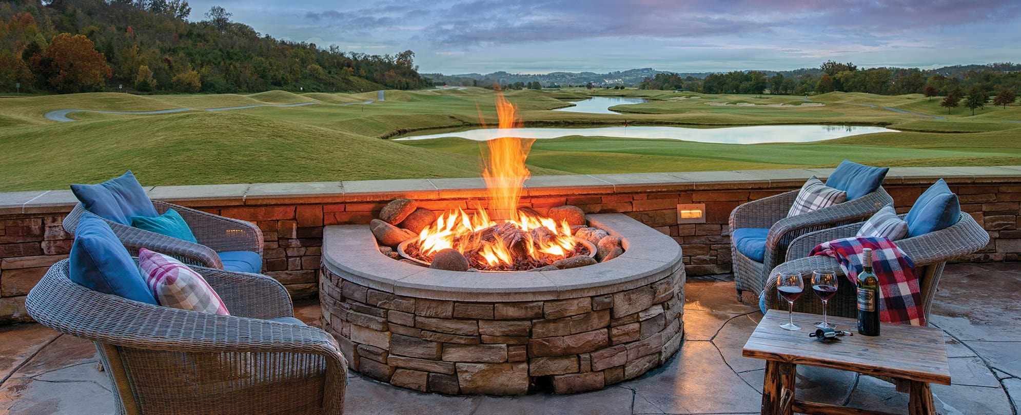 Four comfortable seats around a  firepit with a table that has a bottle of wine and two glasses filled with red wine, overlooking the golf course on the Club Wyndham property. 