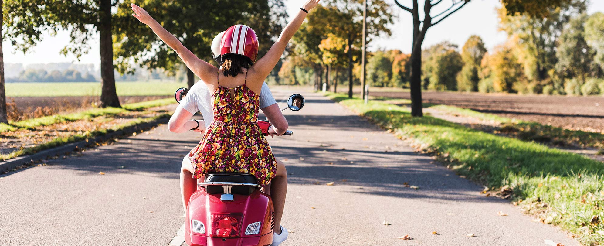 A woman on the back of a motorcyle raises her hands in the air while the man in front of her drives along a scenic tree-lined road