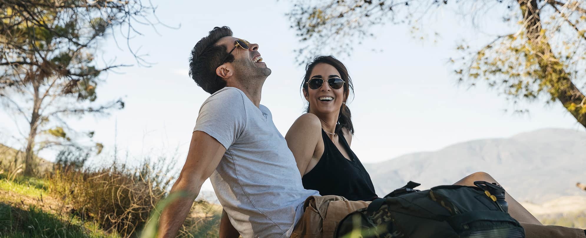 A man and woman sitting on a grassy hillside with mountains in the background, they are both smiling and wearing sunglasses.
