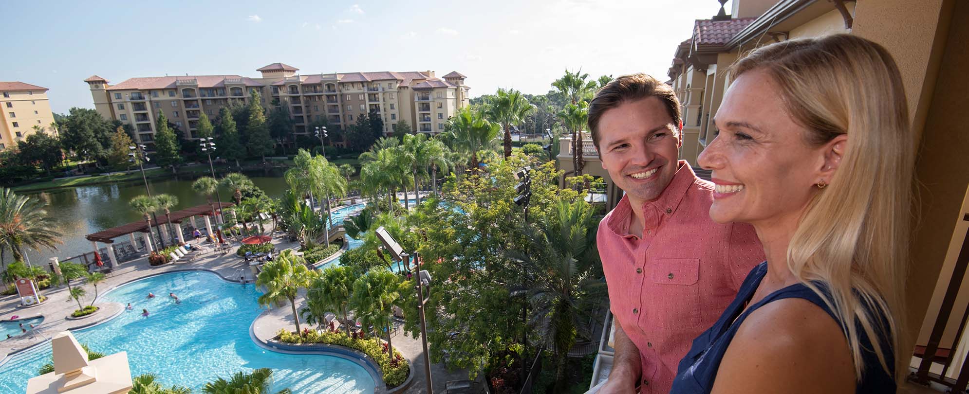 A smiling couple stands on a suite balcony overlooking the pool and lazy river at Club Wyndham Bonnet Creek in Orlando, FL.