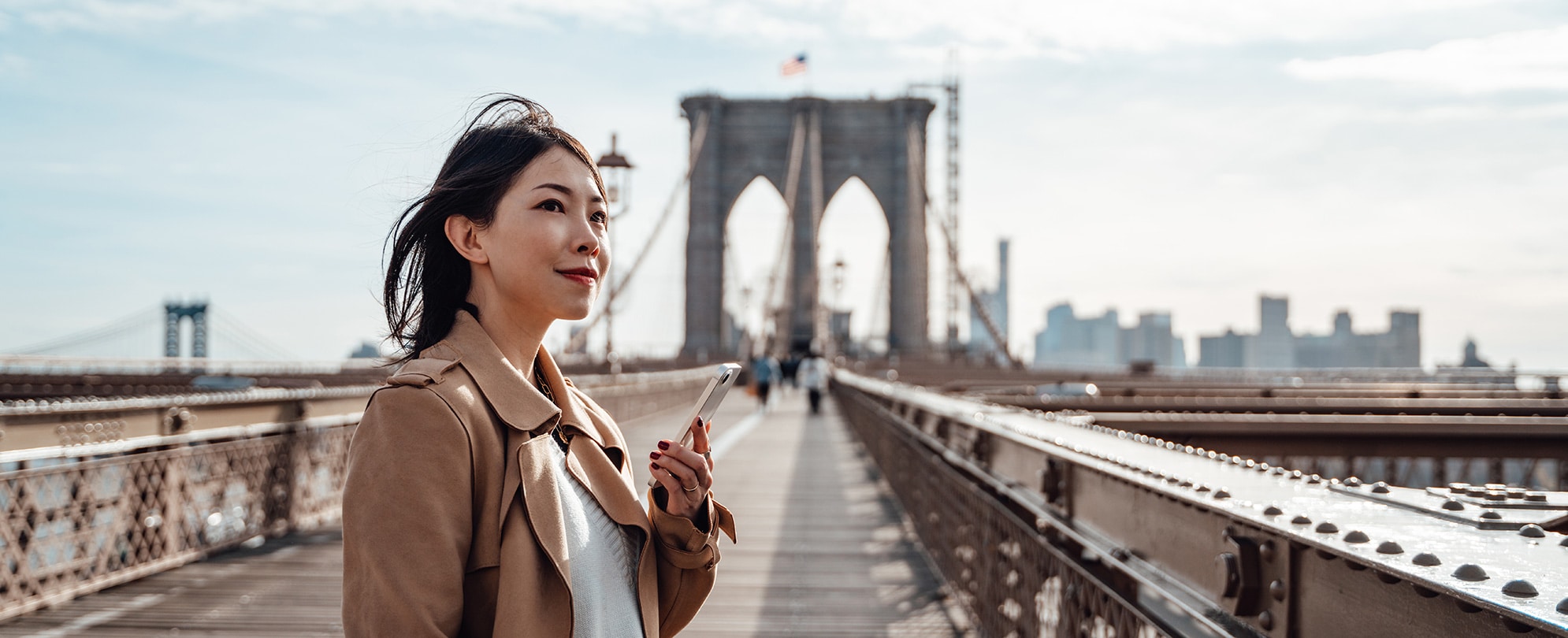 A woman stands on the Brooklyn Bridge looking out over the city and holding her cell phone about to make a phone call.