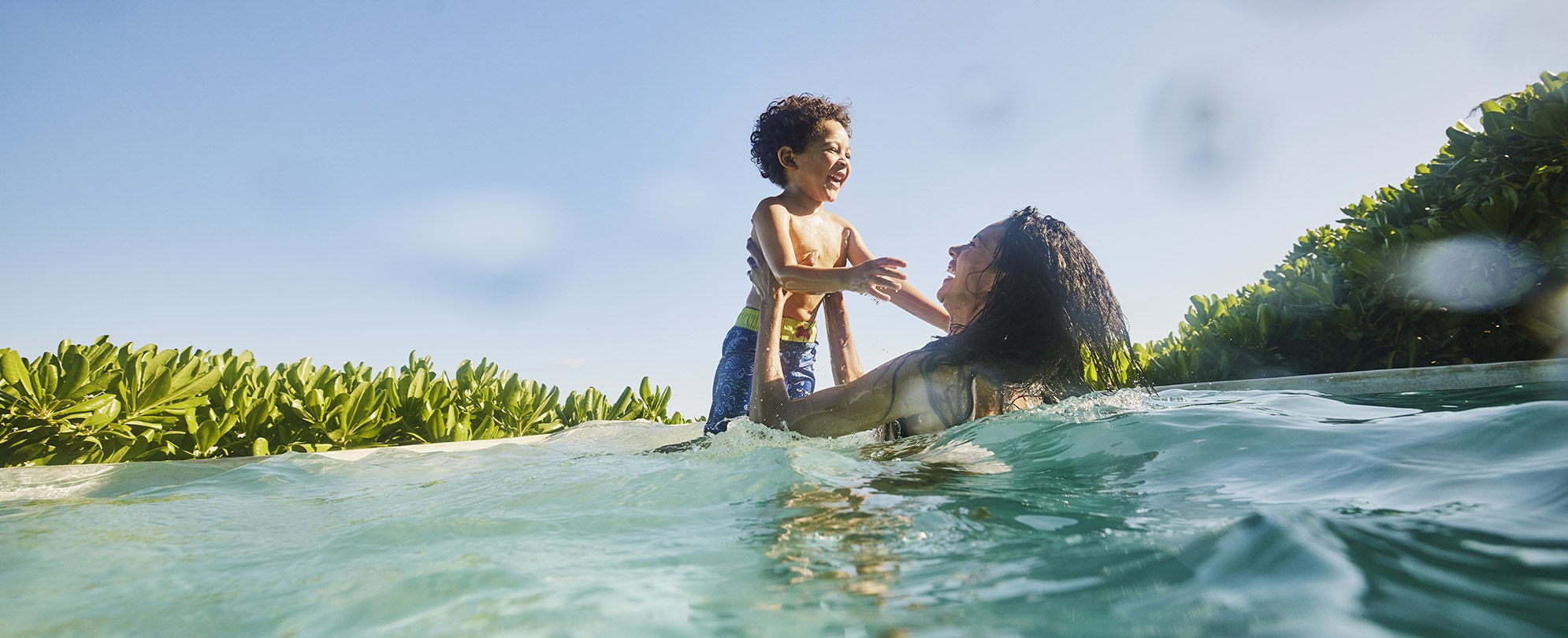 A woman holds a little boy up out of the water as they both laugh while swimming in the ocean.