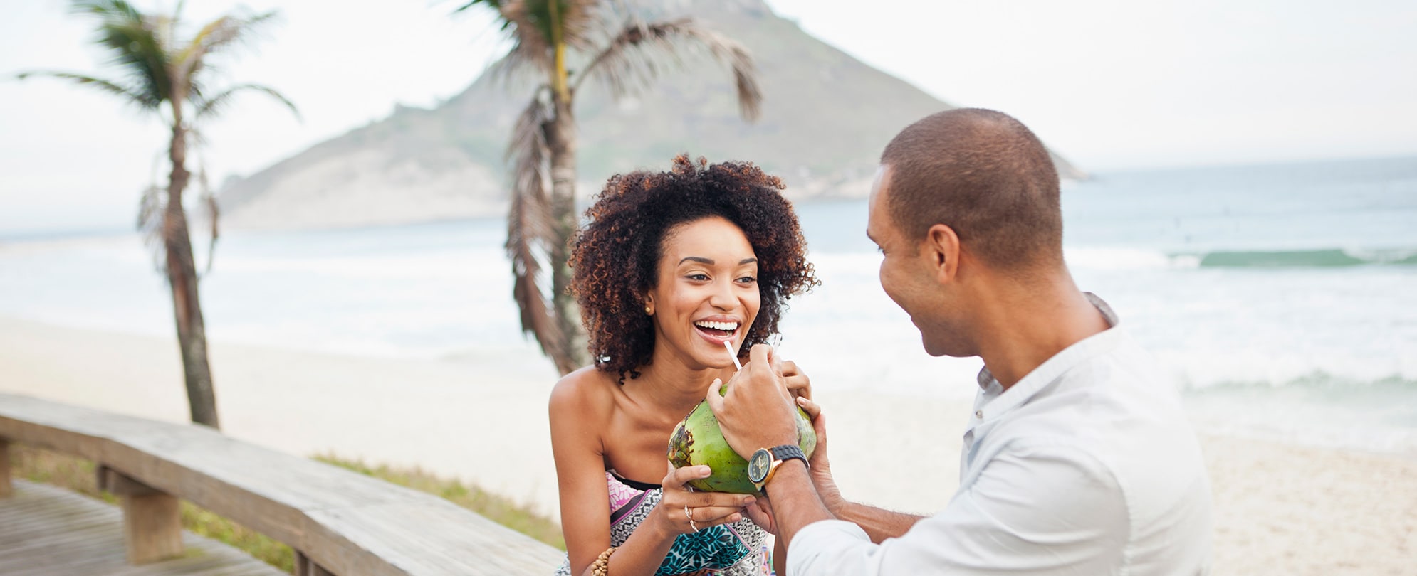 A man offers a woman a drink from a straw sticking out of a coconut as they sit on a circular wood bench on the coast of Hawaii with palms trees and an island volcano in the background.