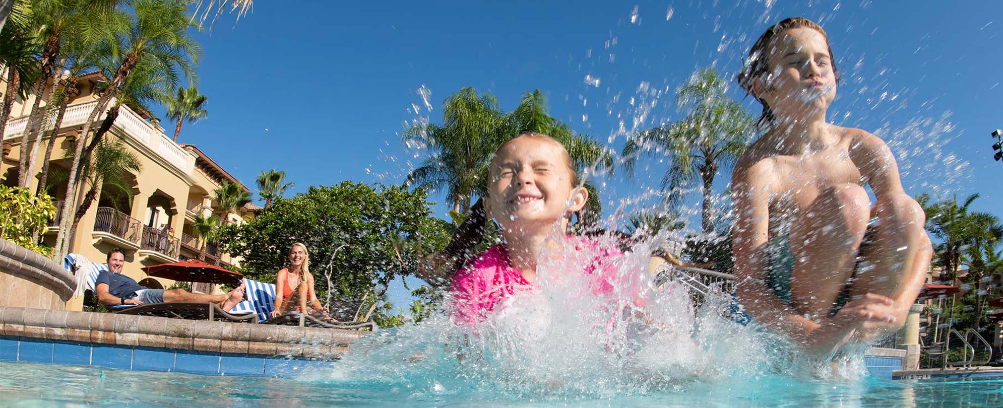 Smiling parents watch as their son and daughter jump and splash in the pool of their Club Wyndham timeshare resort.