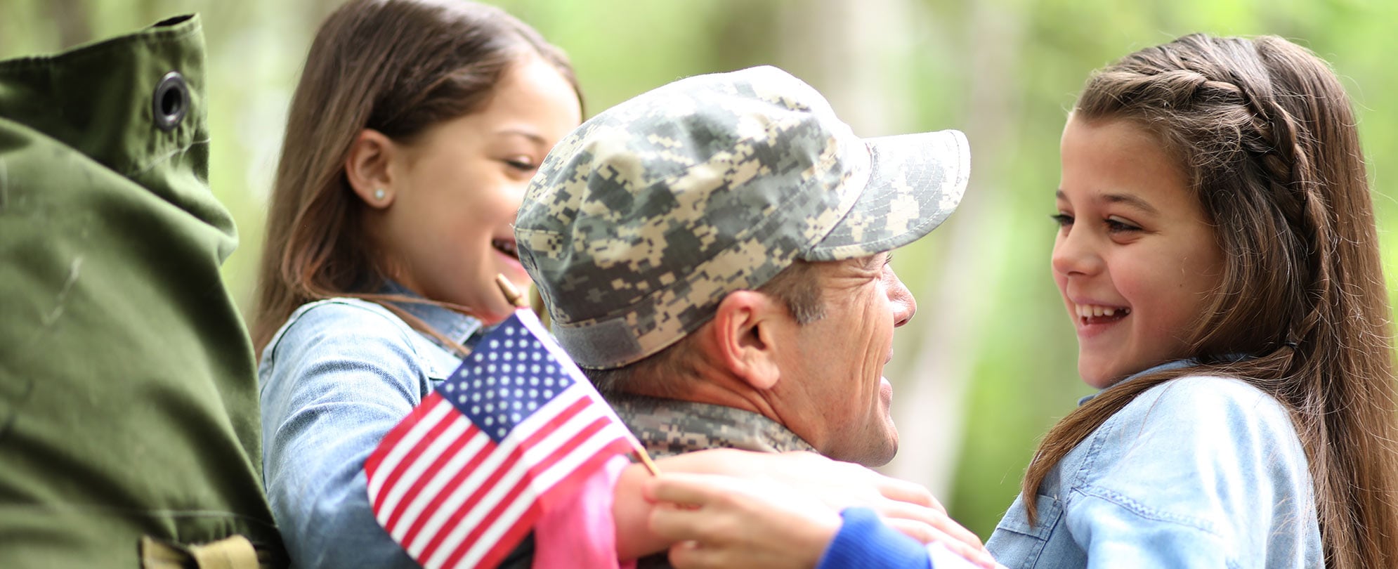 A man in military uniform smiles as he hugs two twin girls, one holding a small American flag.