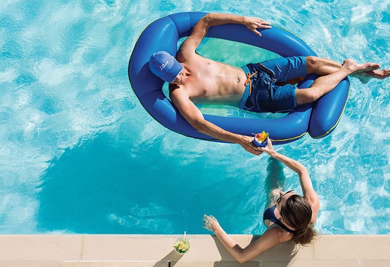 A woman standing in a resort pool is handing a drink to a man laying on a blue pool float. 