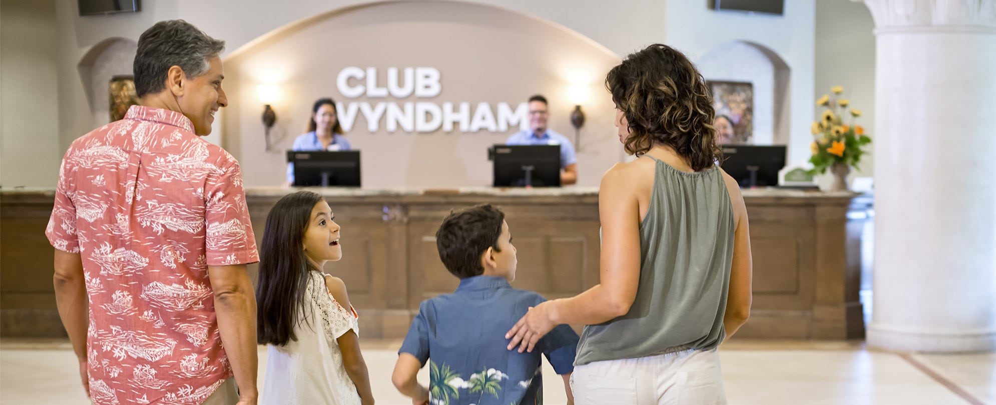 An excited family of four walking through the lobby towards the front desk of a Club Wyndham resort.