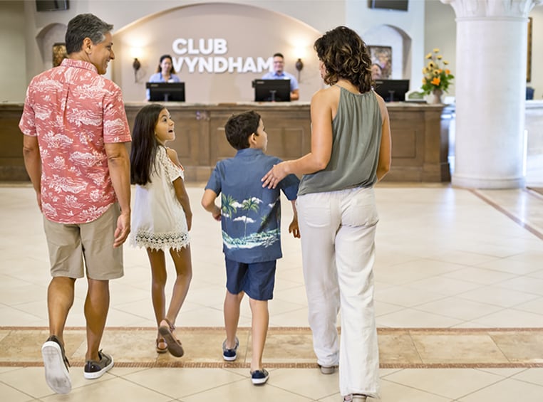 An excited family of four walking through the lobby towards the front desk of a Club Wyndham resort.