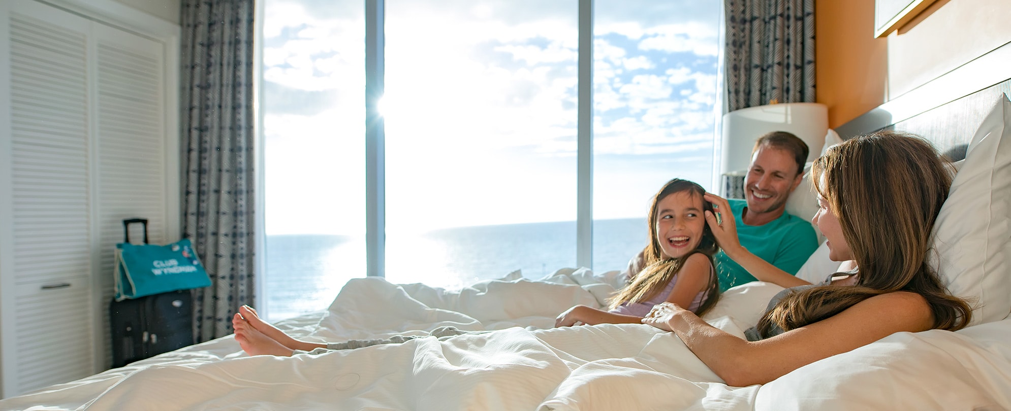 A family of three sits in their bed in a Club Wyndham suite with a beautiful view of the ocean outside