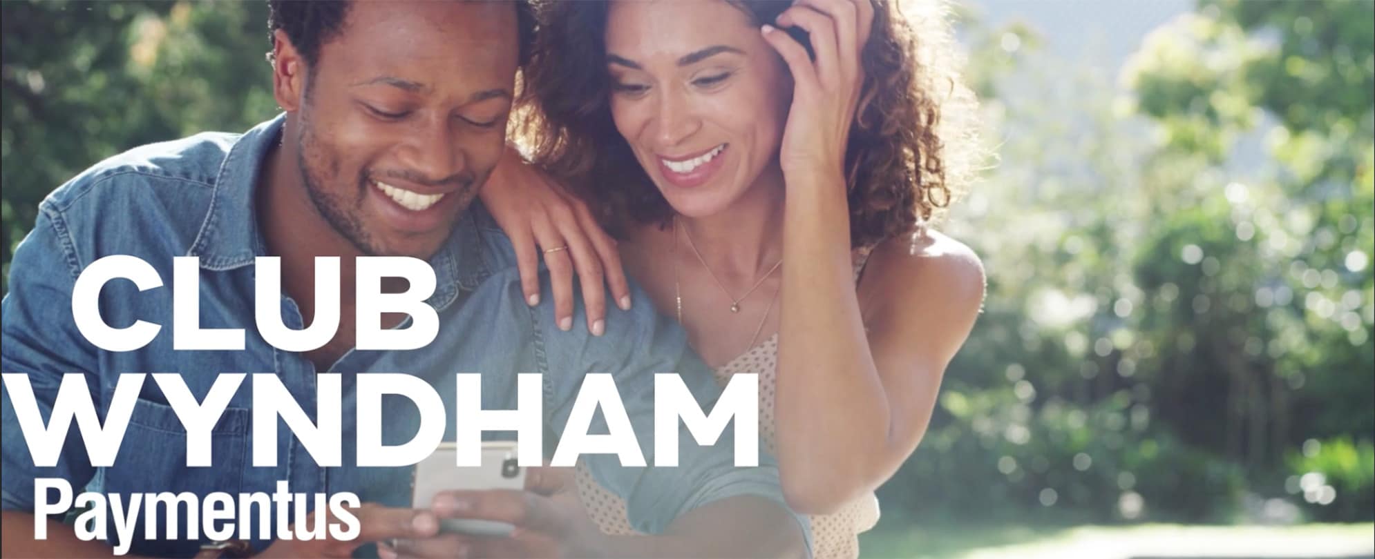 A woman looks over her husband's shoulder as they both smile looking at his cell phone with the Club Wyndham and Paymentus logos overlayed on the image.