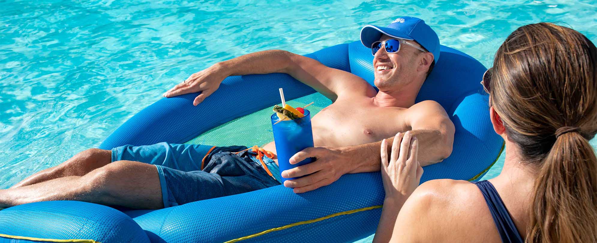Woman standing in the resort pool next to a Man wearing a Club Wyndham hat, who is laying in a pool float, and holding a cocktail. 