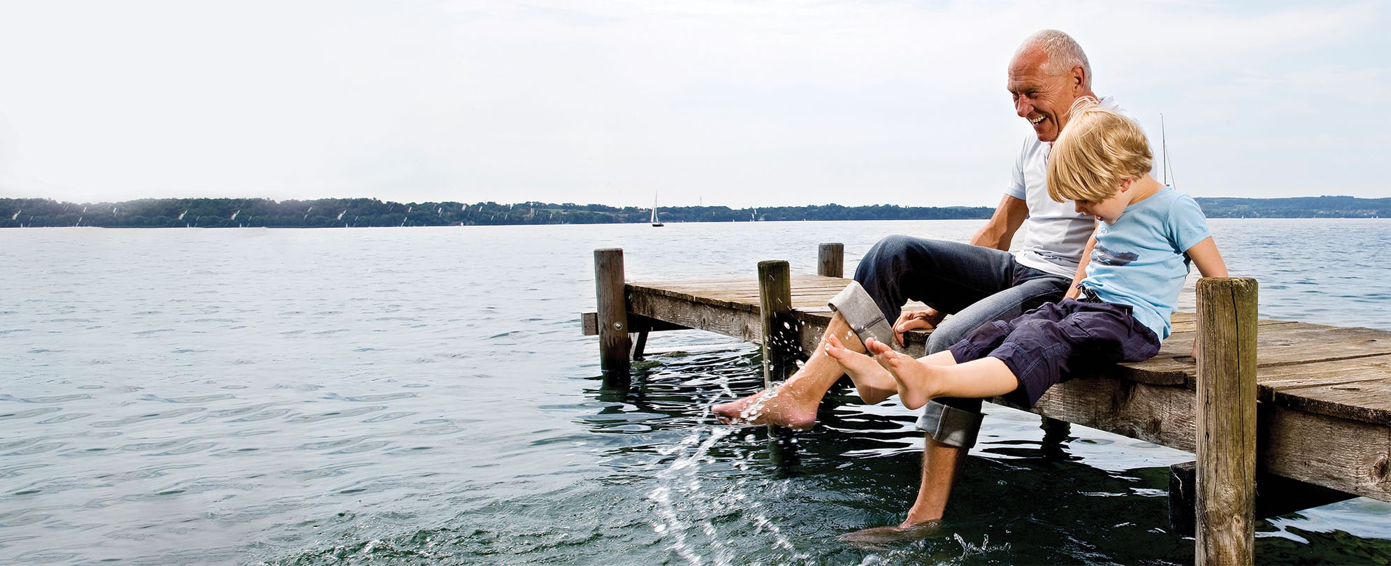 A grandpa and grandson sit on the edge of a dock while splashing the water with their feet