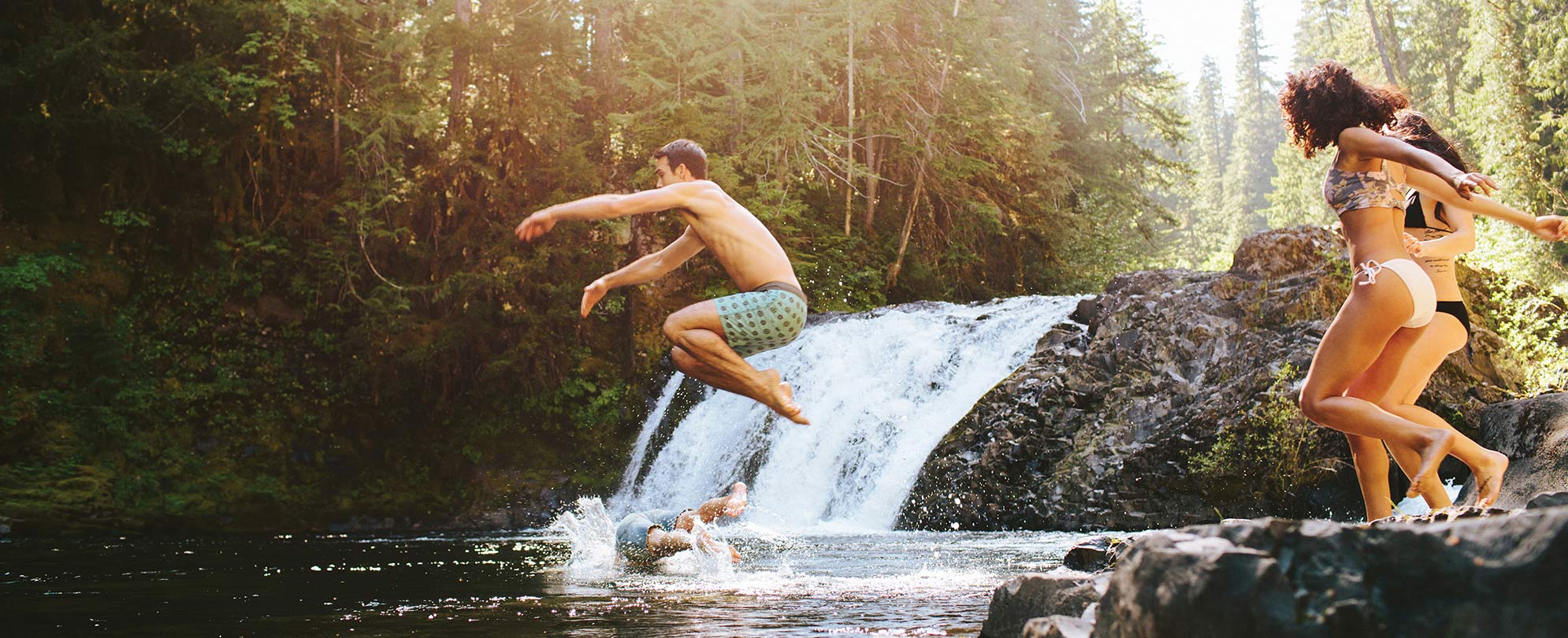 A group of friends jump into a swimming spot by a waterfall.