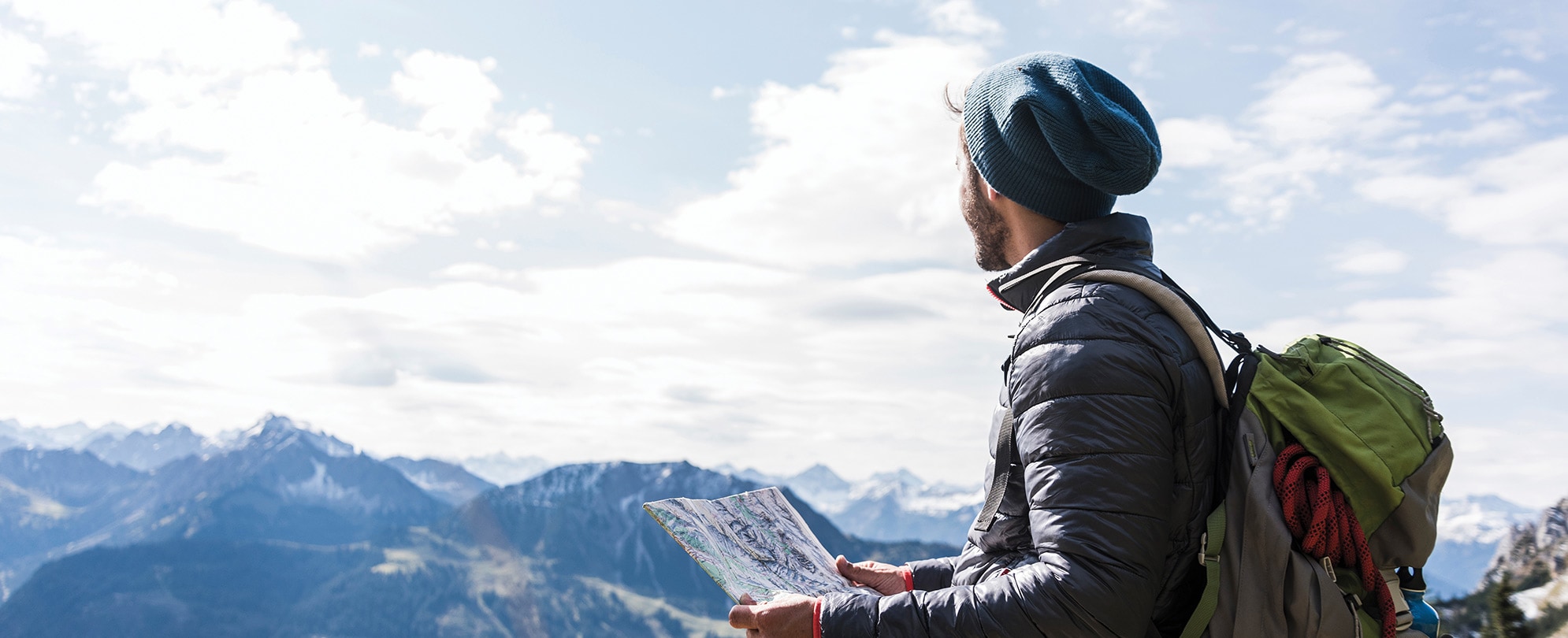 A hiker wearing a coat, backpack, and beanie looks out over a mountain range. 