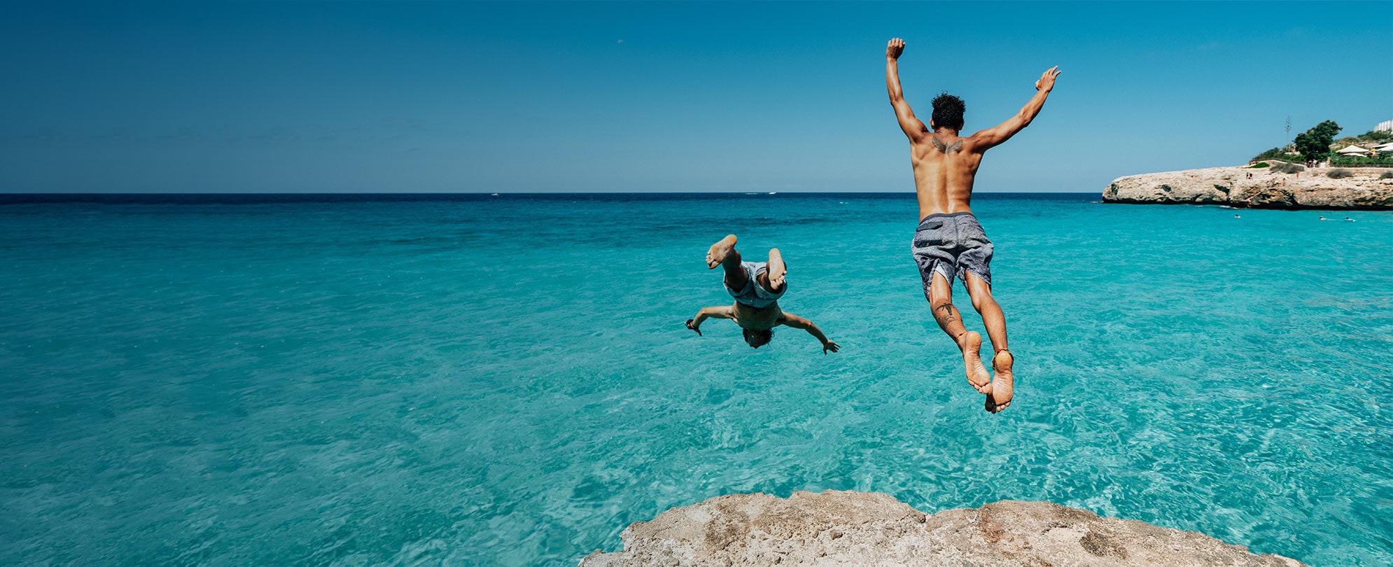 Two men jumping off a rock into the ocean. 
