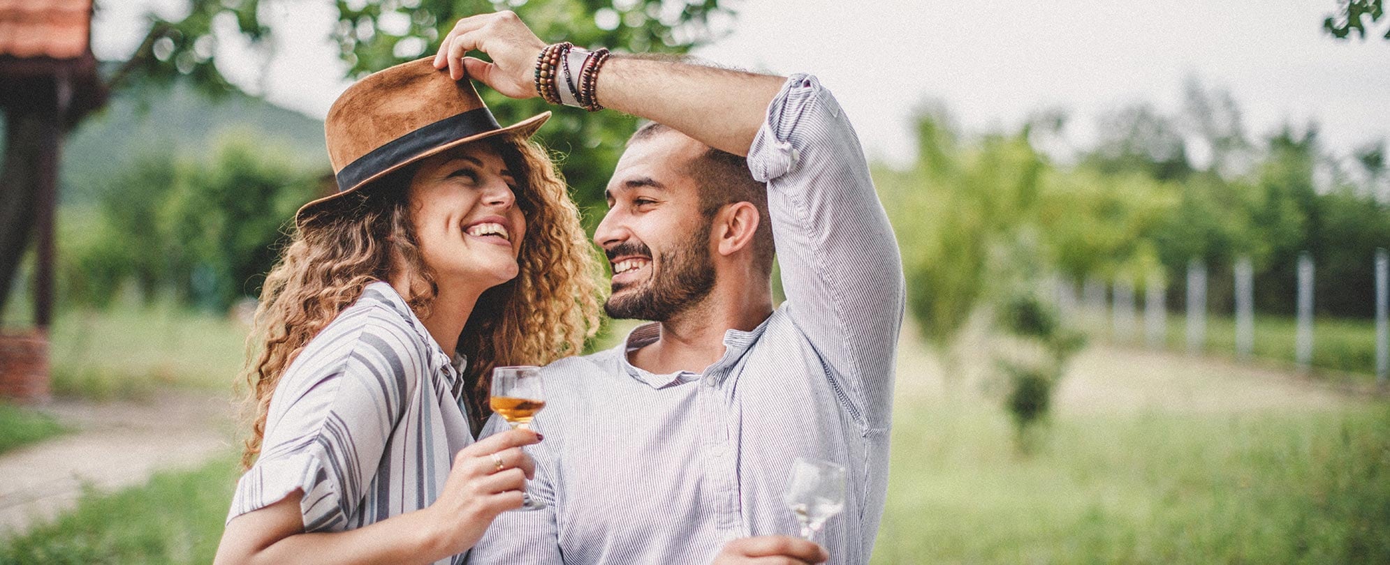 A smiling couple holding wine glasses outside and the man is playfully putting a hat on the woman’s head