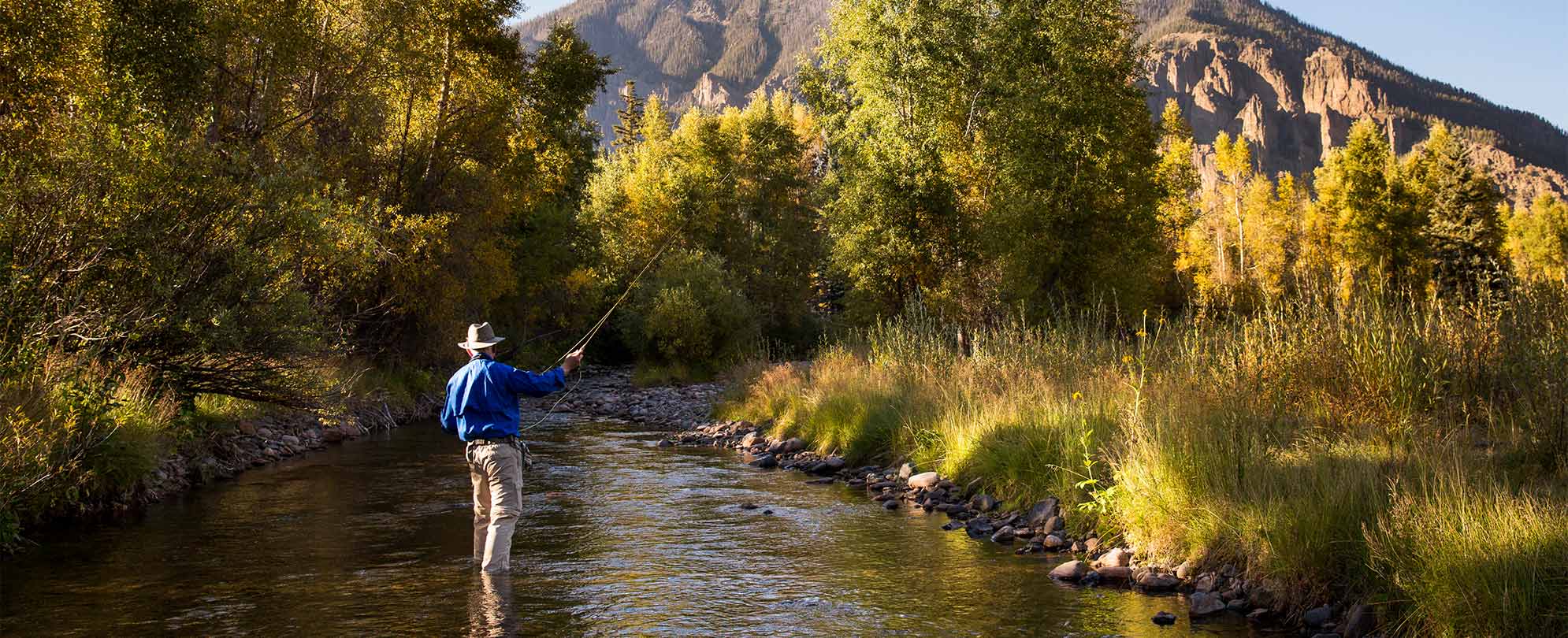 A man stands fishing in a stream, the mountains of Pagosa Springs, Colorado in the distance.